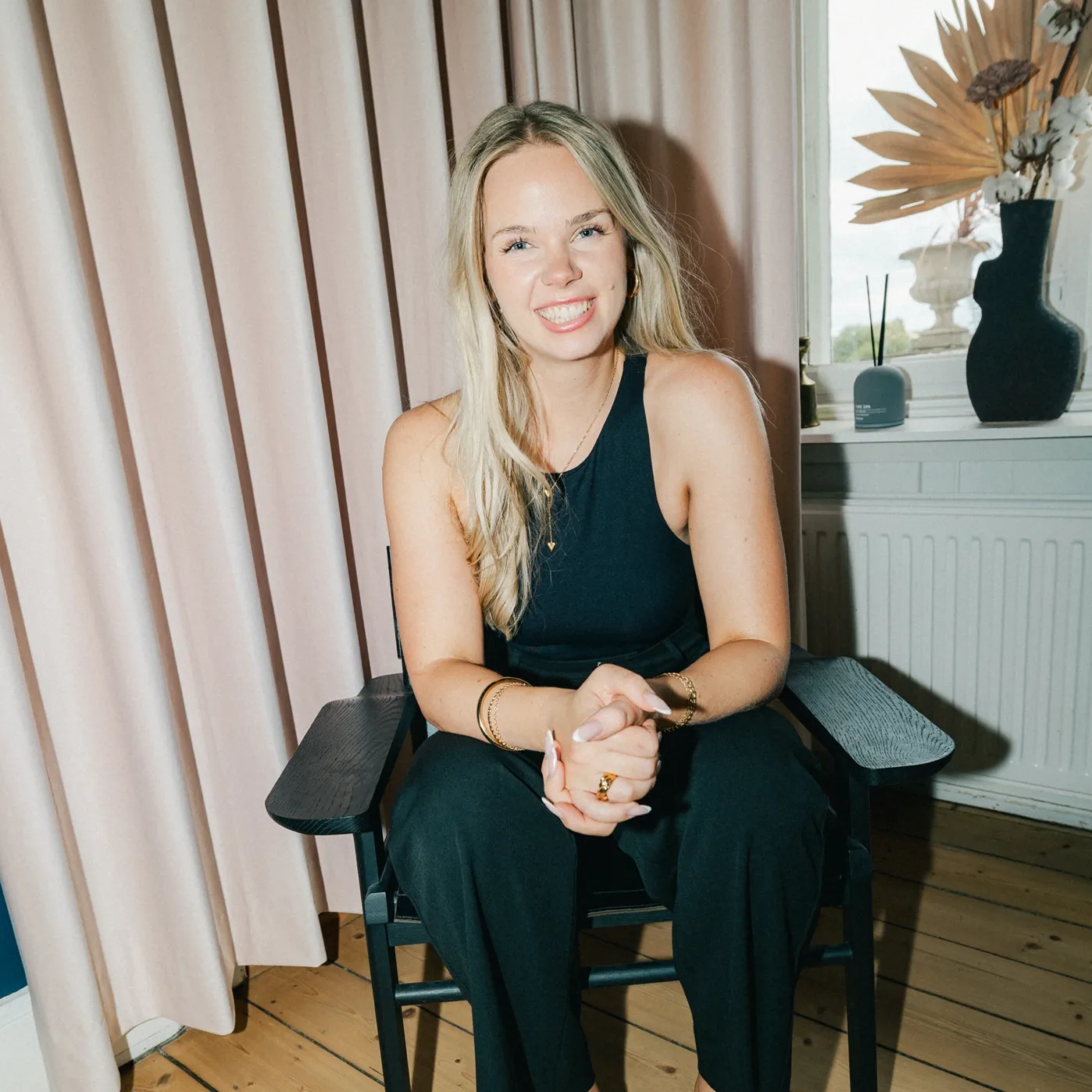 Medarbejder sidder smilende med foldede hænder på en sort stol foran et lyst gardin