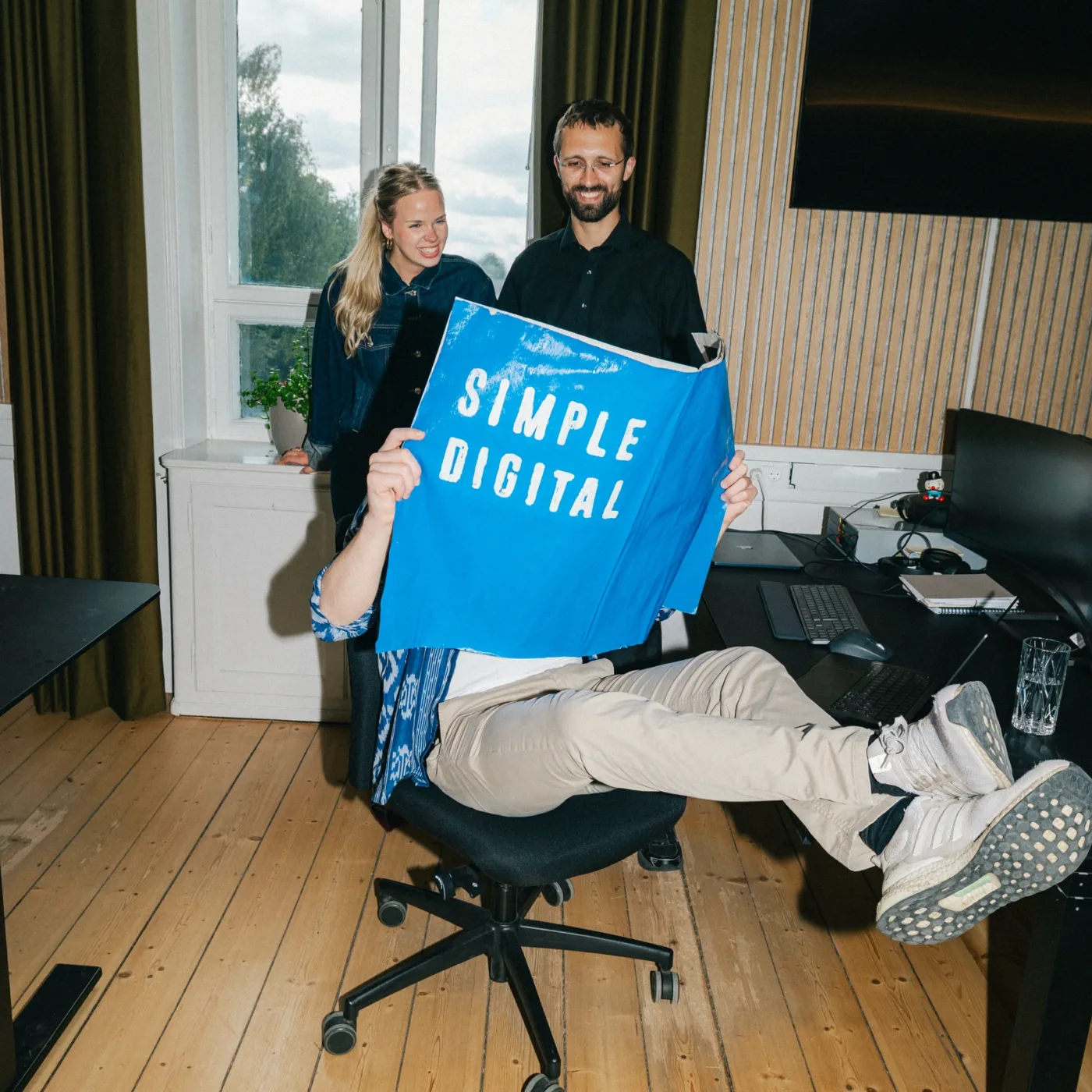 Kolleger griner sammen om en blå plakat med teksten Simple Digital på kontoret
