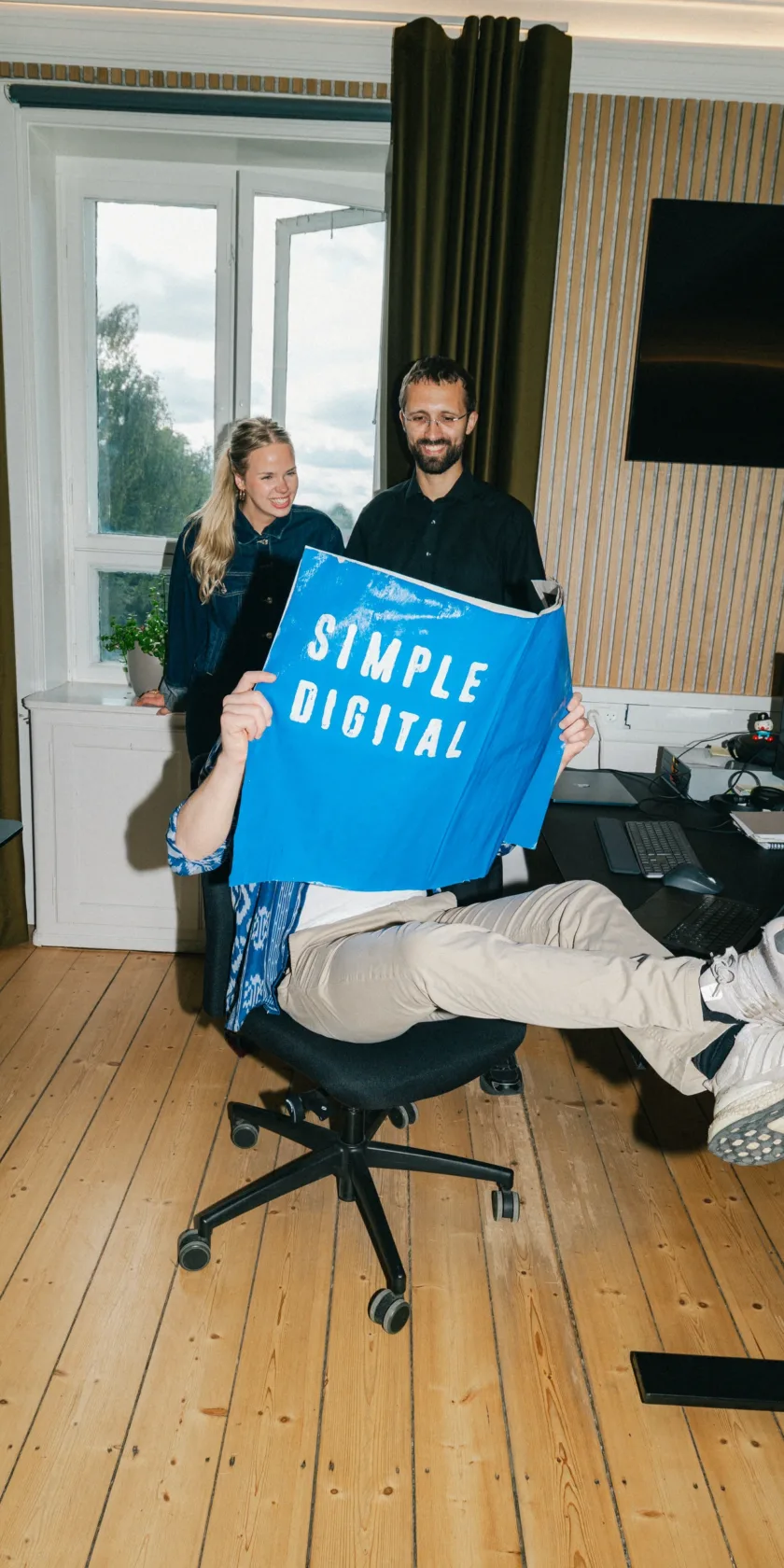 Kolleger holder en blå Simple Digital plakat op og griner sammen på kontoret